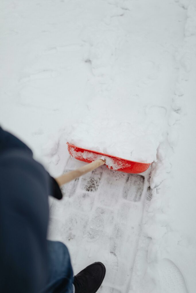 pexels-photo-6952455-6952455 Person shoveling snow from a cobblestone sidewalk with a red shovel in winter.